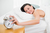 Smiling woman in bed extending hand to alarm clock