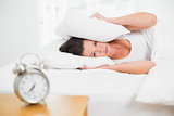 Woman covering ears with pillow and alarm clock on side table