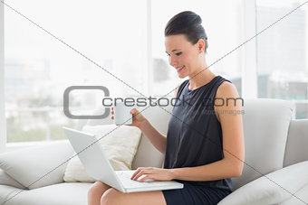 Beautiful well dressed woman using laptop while having coffee on sofa