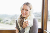 Smiling woman wearing earmuff with coffee cup against window