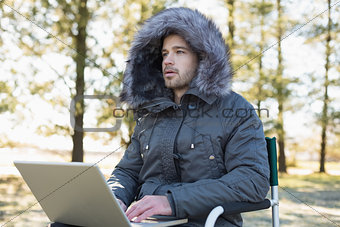 Young man in fur hood jacket using laptop in forest