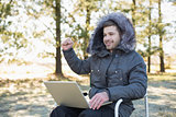 Cheerful man in fur hood jacket using laptop in the forest