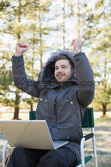 Cheerful man in fur hood jacket with laptop clenching fists in forest