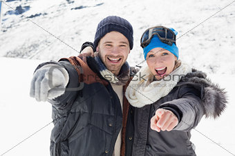 Cheerful couple in jackets pointing at camera on snow covered landscape