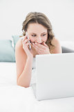 Smiling woman using cellphone and laptop in bed
