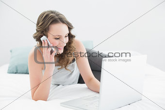 Smiling casual woman using cellphone and laptop in bed