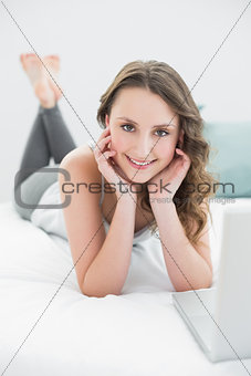Smiling casual young brunette with laptop in bed
