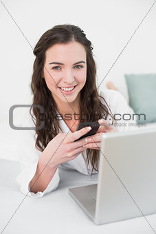 Smiling casual woman with cellphone and laptop in bed