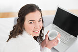 Casual brunette with coffee cup and laptop in bed