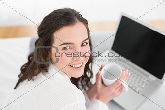 Casual brunette with coffee cup and laptop in bed