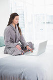 Serious brunette in bathrobe using laptop in bed