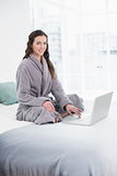Smiling brunette in bathrobe using laptop in bed
