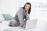 Smiling brunette in bathrobe using laptop in bed