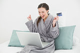 Excited woman in bathrobe doing online shopping in bed