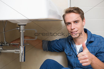 Handsome plumber gesturing thumbs up besides washbasin drain