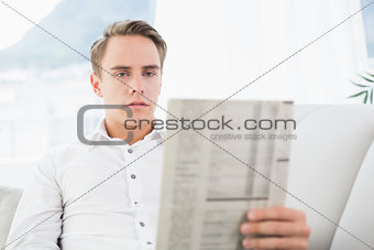 Concentrated relaxed man reading newspaper on sofa