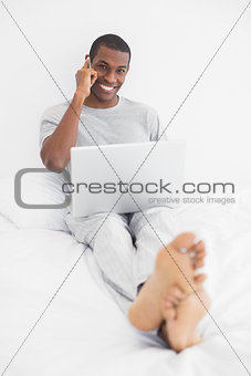 Afro young man using cellphone and laptop in bed