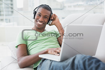 Smiling young Afro man with headphones using laptop on sofa
