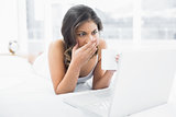 Shocked woman drinking coffee while using laptop in bed