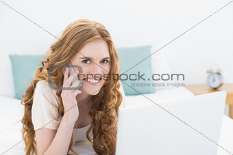 Portrait of smiling casual woman using laptop and cellphone in bed