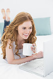 Smiling blond with coffee cup using laptop in bed