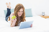 Blond using tablet PC while holding an apple in bed