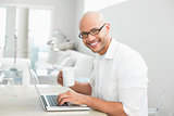 Smiling casual man with coffee cup using laptop at home