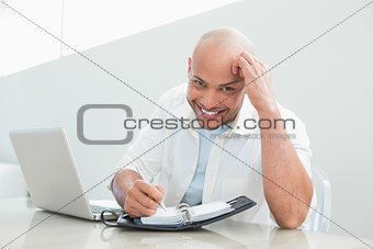 Casual smiling man with laptop writing in diary at home