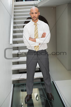 Serious businessman standing with arms crossed against staircase