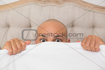 Shocked bald man covering face with sheet in bed