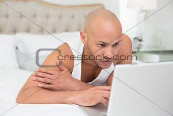 Serious casual bald man using laptop in bed