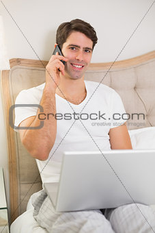 Casual smiling man using cellphone and laptop in bed