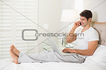 Casual man using cellphone and laptop in bed