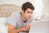 Serious casual young man using laptop in bed