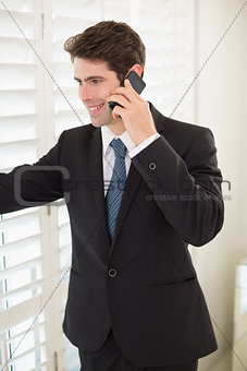 Smiling businessman peeking through blinds while on call