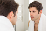 Serious young man looking at self in bathroom mirror