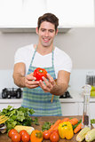 Smiling man holding out bell pepper with vegetables in kitchen