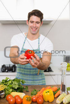 Smiling man holding out bell pepper with vegetables in kitchen