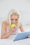 Casual blond eating an apple while using tablet PC in bed