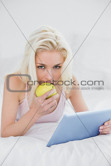 Casual young blond with tablet PC eating an apple in bed