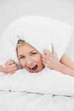 Angry young woman covering ears with pillow in bed
