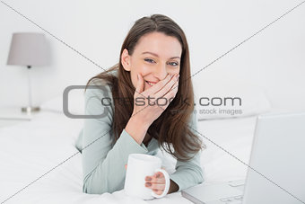 Cheerful casual woman with laptop and cup in bed