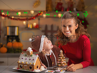 Smiling mother and baby with christmas cookie house in kitchen