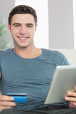 Cheerful handsome man relaxing on couch using tablet shopping online