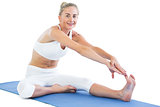 Toned smiling blonde sitting on exercise mat stretching right leg