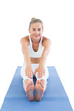 Toned smiling blonde sitting on exercise mat stretching legs