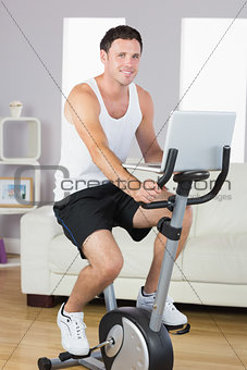 Smiling sporty man exercising on bike and holding laptop