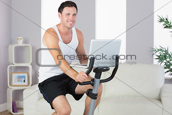 Cheerful sporty man exercising on bike and holding laptop
