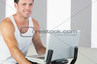Handsome sporty man exercising on bike and holding laptop