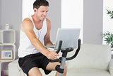 Sporty man with earphones exercising on bike and holding laptop